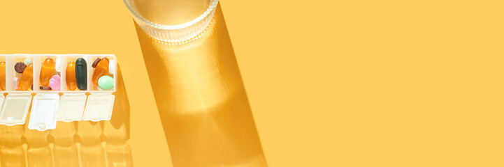 Assorted pills in weekly organizer and glass of water on bright yellow background.