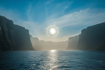 A glowing sun halo forming above a fjord surrounded by cliffs