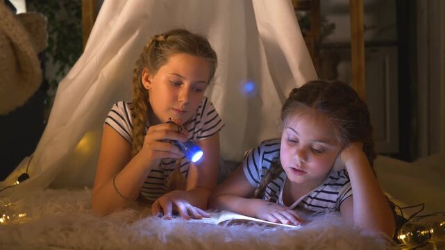 Girls read under blanket with flashlight while sister leans close in cozy indoor tent lit by fairy light string as child and girl follow book page during bedtime reading session with soft warm glow