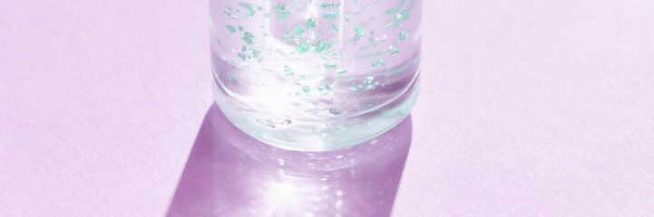 Transparent gel bottle with green particles on pastel purple background casting shadow.