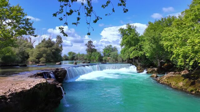 Bright vivid waterfall in Manavgat national park splashing azure water over rocky cliffs with surrounding forest glowing green under blue sky during sunny summer day, slow motion