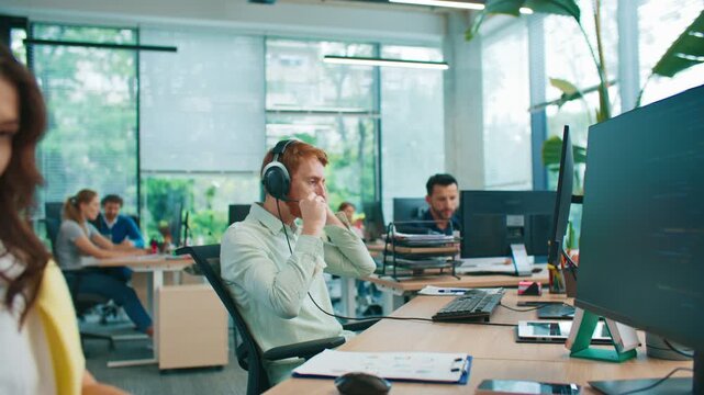 Red-haired man sitting at desk while putting on headset. Preparing for online meeting in open office. Coworkers working and talking in background. Team focusing on communication and collaboration.