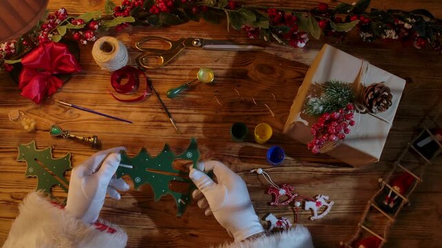 Top view of Santa's gloved hands creating festive decorations with paint, ribbons, and scissors next to a wrapped gift on a rustic wooden table.