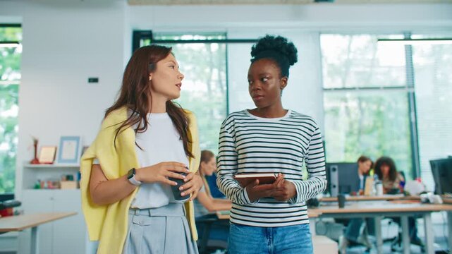Two women walking side by side in bright office space talking about work. One holding coffee cup other carrying notebook. Team visible in background working at desks. Daylight workplace conversation.