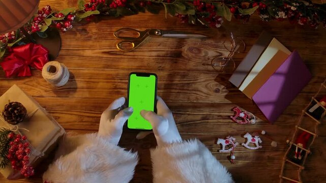Top view of Santa Claus's gloved hands typing on a smartphone with a green chroma key screen on a rustic wooden table. Preparing for New Year's and Christmas celebrations.