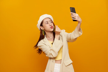 Beautiful young Asian woman shows mini heart fingers taking selfie with smartphone isolated on yellow background. expressing playful confidence and modern digital lifestyle