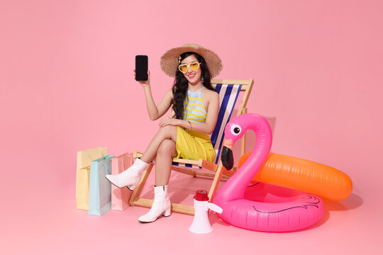 Asian woman sitting on beach chair holding smartphone blank screen and shopping bags with inflatable flamingo symbolizing summer lifestyle online shopping and digital vacation concept