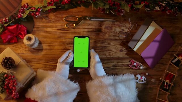 Top view of Santa Claus's gloved hands swiping on a smartphone with a green chroma key screen on a rustic wooden table. Preparing for New Year's and Christmas celebrations.