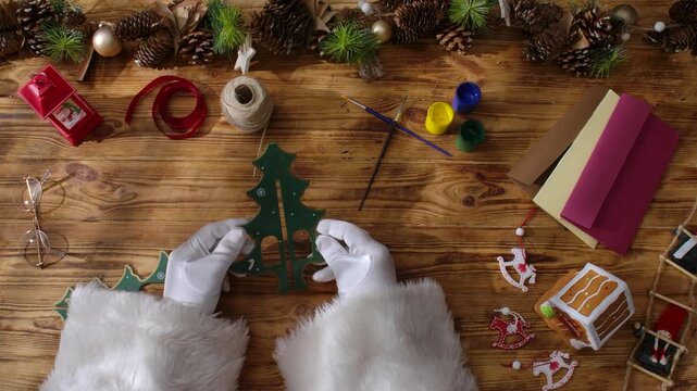 Top view of Santa's gloved hands creating festive decorations with paint, ribbons, and scissors next to a wrapped gift on a rustic wooden table.