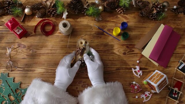 Top view of Santa Claus's gloved hands arranging Christmas tree decorations in the shape of various birds on a rustic wooden table. Preparations for New Year's and Christmas celebrations.
