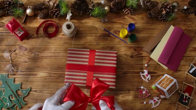 Top view of Santa Claus's gloved hands creating festive gifts with his own hands on a rustic wooden table. Preparing for New Year's and Christmas celebrations.