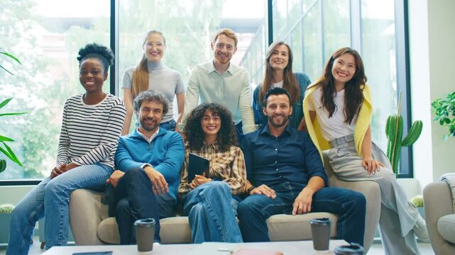 Group of men and women posing together on sofa in bright modern office smiling and laughing. Happy diverse coworkers sitting and standing for team photo. Friendly workplace environment.