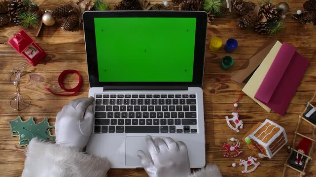 Top view of Santa's gloved hands works on a laptop keyboard with a green chroma key screen on a rustic wooden table. Preparing for New Year's and Christmas celebrations.