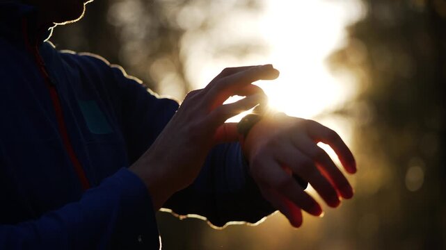 Close-up of a silhouetted athlete using a smart watch to track performance during a training run in a park. The morning sun creates a beautiful lens flare, illuminating the hands