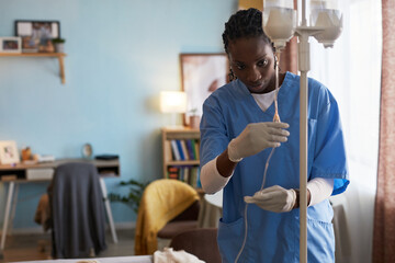 Black woman young adult nurse adjusting intravenous drip in medical home care setting, wearing gloves and scrubs, focusing on patient, standing near IV stand in clinical environment