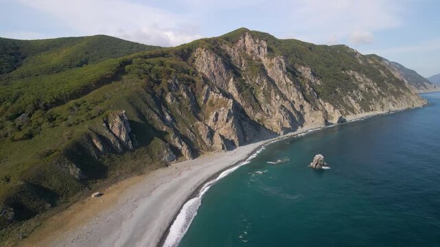 Aerial drone footage of the bay. A long pebble beach, turquoise waves, dramatic sea stacks and steep rocky cliffs along the wild Pacific coastline.