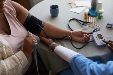 Elderly Black woman with disability having blood pressure measured by healthcare worker using digital monitor, medication bottles and stethoscope visible on table