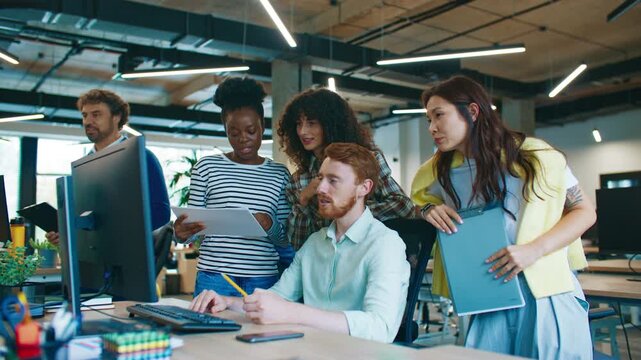 Man sitting at desk in modern office answering questions while young coworkers standing nearby learning and discussing. Interns asking for advice and taking notes. Productive teamwork atmosphere.