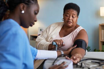Middle aged Black woman receiving blood pressure measurement from young adult Black female healthcare worker in medical setting, patient looking down while nurse operating device