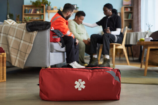 Black female paramedic comforting middle aged Black woman sitting on couch while Caucasian male paramedic supporting during emergency response, red medical bag in foreground