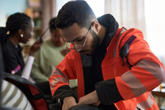 Young adult Caucasian man wearing emergency responder uniform preparing medical equipment while sitting indoors, with two Black women in background engaging in conversation