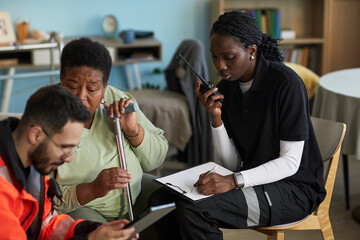 Middle aged Black woman holding cane sitting next to young female nurse using walkie talkie and...