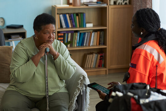 Middle aged Black woman sitting on sofa holding cane with both hands, looking at female paramedic with tablet, receiving medical consultation in home setting