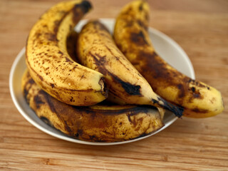A bunch of overripe bananas with brown spots resting on a white plate on a wooden table. Shallow depth of field. Close up.
