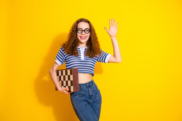 Young woman with curly hair waves hello while holding a chessboard against a bright yellow...