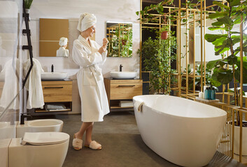 Full length portrait of cheerful attractive young woman in white bathrobe and towel on head relaxing in modern bathroom interior enjoying morning time at home. Beauty, wellness and hygiene concept.