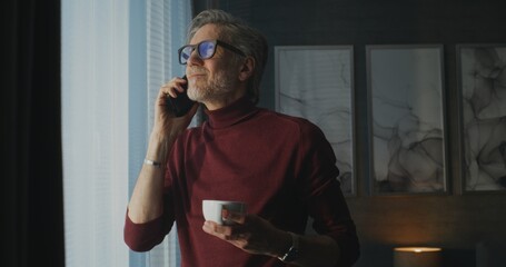 Stylish Mature Man in Red Turtleneck and Glasses Stands By Panoramic Window in Bright Hotel Room, Sipping Coffee While Talking on Phone. Concept Morning Routine, Business Travel, Remote Communication.