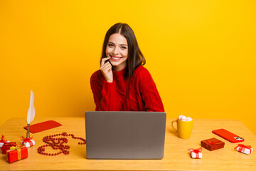 Cheerful young woman in a red sweater works on a laptop at a festive desk with gifts mug and...