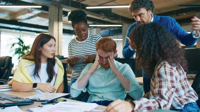 Team gathered around stressed male coworker holding head in hands during discussion. Colleagues talking and gesturing while trying to solve problem together at workplace in open office setting.