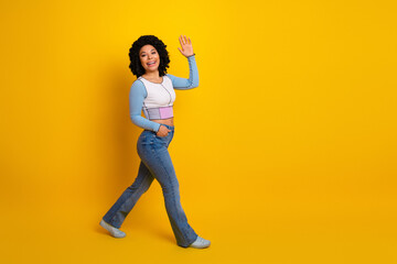 Smiling young woman waving against a yellow background, dressed in casual fashion with a cheerful expression