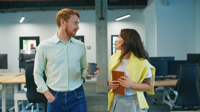 Smiling coworkers spending break together. Male with beard holding coffee cup and woman carrying notebook talking cheerfully. Sharing friendly moment between tasks. Standing near desks in office area.