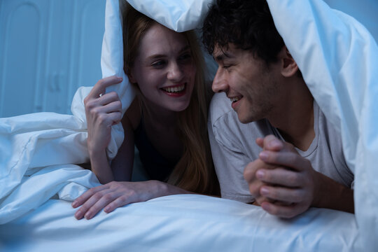 Happy young couple lying in bed under blanket, smiling and talking at night. Intimate romantic moment, cozy atmosphere, relationship bonding, bedtime, and night routine concept. couple, love, bed. - Powered by Adobe