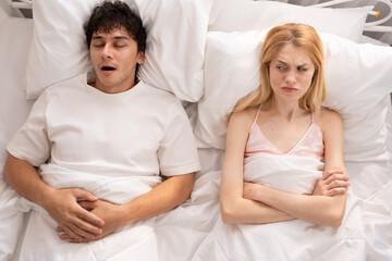 Frustrated woman sitting in bed covering her ears while her partner snores loudly, sleep disturbance, relationship issues, and frustration in a shared bedroom setting. snoring, couple, bed, sleep.