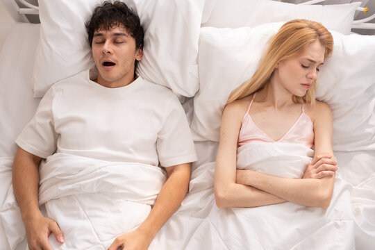 Frustrated woman sitting in bed covering her ears while her partner snores loudly, sleep disturbance, relationship issues, and frustration in a shared bedroom setting. snoring, couple, bed, sleep. - Powered by Adobe