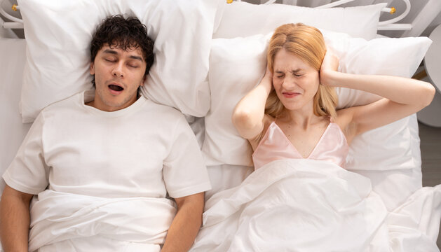 Frustrated woman sitting in bed covering her ears while her partner snores loudly, sleep disturbance, relationship issues, and frustration in a shared bedroom setting. snoring, couple, bed, sleep.