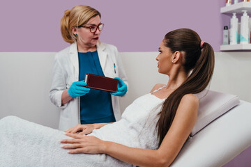 Cosmetologist in blue gloves and lab coat presenting various skincare products to a smiling female client lying on a treatment bed in a modern beauty clinic