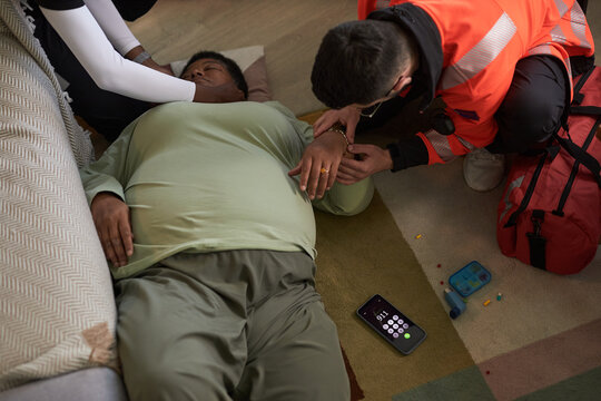 Middle aged Black woman lying on floor receiving emergency medical assistance from paramedic and another , smartphone with emergency call screen and pill organizer visible nearby