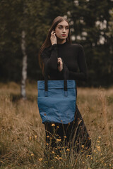 A woman in a long black dress posing in a natural autumn landscape, presenting a blue, ecological shoulder bag, in a balanced fashion style