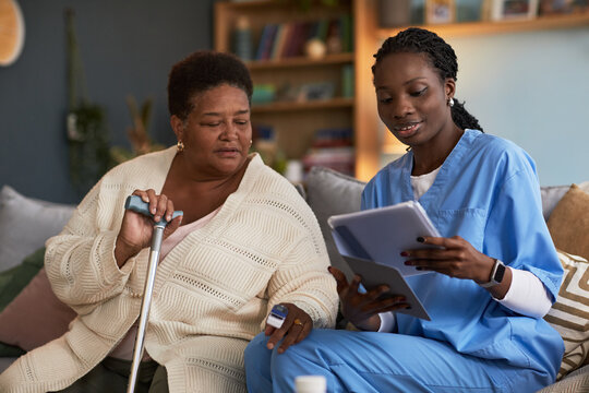 Middle aged Black woman holding walking cane and medical device sitting next to young adult Black woman nurse showing digital tablet, both focusing on healthcare discussion