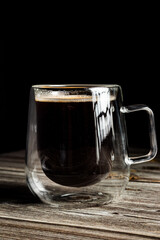 Black coffee in a double-walled glass cup with strong reflections, placed on rustic wooden surface.