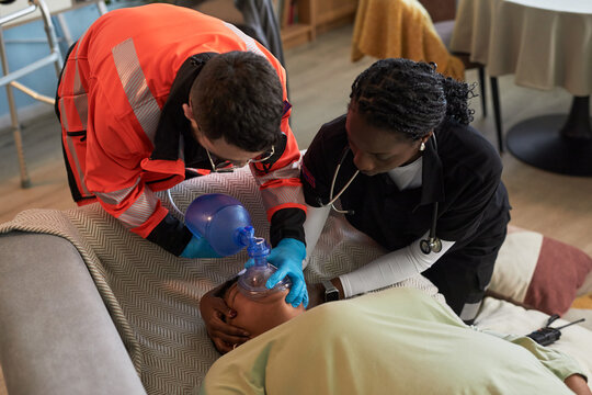 Black woman and Caucasian man paramedic performing emergency resuscitation on unconscious adult using manual resuscitator and monitoring vital signs in home setting
