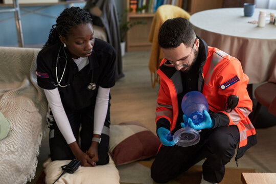 Black woman young adult paramedic sitting on floor observing Caucasian man young adult paramedic preparing manual resuscitator during emergency medical scenario in home setting