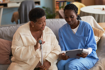 Senior Black woman with disability sitting on sofa holding cane, listening to young adult Black female nurse in medical scrubs showing information on digital tablet