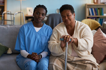 Portrait of young adult Black woman caregiver sitting beside senior Black woman with disability holding cane, both looking at camera, showing support and companionship on sofa during home visit