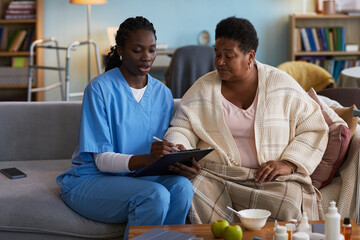 Black young adult woman wearing medical scrubs sitting on sofa showing clipboard to Black middle aged woman wrapped in blanket, both focusing on healthcare discussion in living room