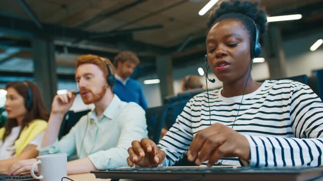 Focused female call center agent wearing striped shirt and headset speaking with client while typing response on keyboard. Customer service representative communicating remotely in office workspace.
